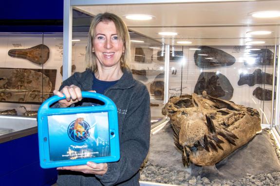 A woman with bob length blonde hair and wearing a blue jumper is holding a tablet with a dinosaur display. She is standing next to a glass cabinet that contains a huge skull of an ancient sea monster